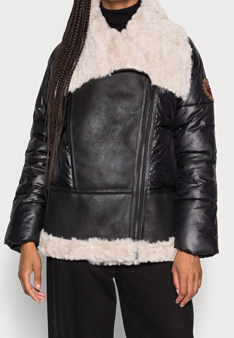 Femme portant une veste en similicuir noir et une doudoune avec un grand col et ourlet en fausse fourrure crème, une fermeture éclair frontale asymétrique et un patch brodé sur la manche.