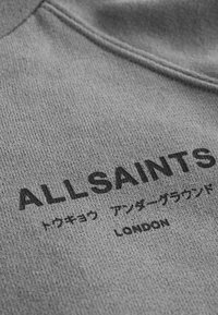 Nahaufnahme eines grauen Stoffes mit schwarzem Text „ALLSAINTS“, japanischen Schriftzeichen und „LONDON“ darauf gedruckt.