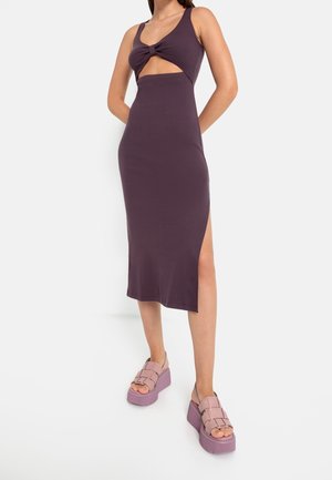 Kvinde iført en ærmeløs mørkelilla midikjole med udskæring foran og knudedetalje, kombineret med lyse lilla platformssandaler.