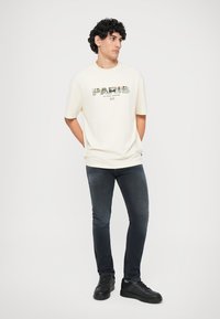 Ung man med lockigt hår som bär en vit t-shirt med texten 'PARIS', mörka åtsittande jeans och svarta sneakers, står med händerna bakom ryggen.