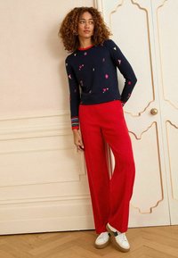 Una donna con i capelli ricci è in piedi contro un muro chiaro, indossando un maglione blu navy con motivi colorati e pantaloni larghi rossi, completati da sneakers.