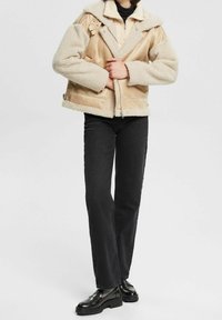 Chaqueta de shearling beige con cuello, mangas texturizadas y secciones de tela lisa en contraste. Combinada con jeans negros de pierna recta y botas negras.