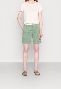 Camiseta beige clara con bolsillo, combinada con pantalones cortos verdes enrollados con un patrón de puntos, complementada con un cinturón beige y sandalias verdes tipo slide.