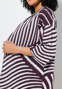 Maglione oversize in un borgogna scuro con strisce zebra bianche, caratterizzato da una trama morbida e maniche ampie. La modella tiene le mani sulla vita.