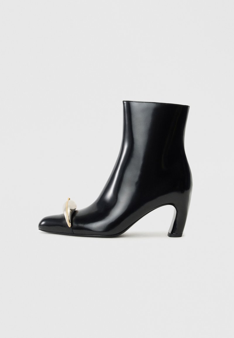 Black ankle boot made of glossy leather, featuring a pointed toe and a decorative gold and silver accent at the front. Block heel design.
