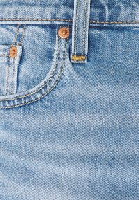 Svetle modré džíny z denimu s měděnými detaily, viditelným šitím a texturovaným povrchem. Obsahují zapínání na knoflík a poutka na pásek.