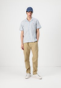 Camisa de manga corta, con botones, de rayas verticales en azul y blanco, combinada con pantalones beiges y zapatillas blancas. También incluye una gorra azul.