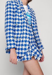 Blazer de houndstooth azul y blanco con acentos de botones dorados de doble botonadura, tejido texturizado y mangas acampanadas. Lleva puesto sobre una camiseta gráfica blanca.