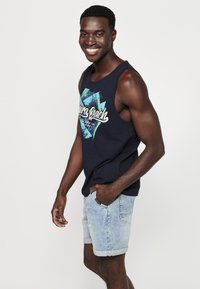 Tumesinine tank top sinise graafilise disainiga, millel on valge tekst, koos heledate teksashortsidega. Modell naeratab ja seisab käed taskutes.