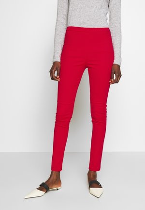 Osoba ubrana w jaskrawo czerwone dopasowane spodnie, szarą bluzkę z długim rękawem oraz białe damskie baleriny z czarnymi paskami, stojąca na białym tle.