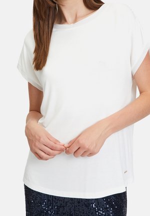 Kobieta w białej koszuli z krótkim rękawem i ciemnej spódnicy z cekinami, ręce delikatnie splecione na talii, długie brązowe włosy na ramieniu.