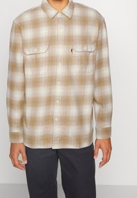 Camicia in flanella a quadri marroni e crema con design a bottoni, due tasche sul petto e maniche lunghe, realizzata in un materiale morbido al tatto.