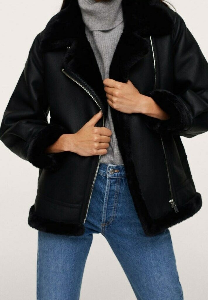 Femme portant une veste d’aviateur noire doublée de fausse fourrure sur un pull à col roulé gris et un jean bleu, tenant les revers de la veste.