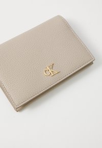 Beige leather wallet with textured surface, featuring a gold "CK" logo and stitched edges. Compact, bifold design for cards and cash.