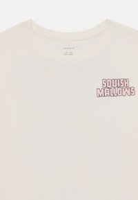 T-shirt a maniche corte con scollo rotondo in cotone bianco sporco. Presenta la scritta rosa "Squish Mallows" sul lato sinistro del petto.