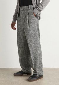 Vida byxor i en strukturerad grått tyg, med veck och sidofickor. Kombinerade med svarta sneakers på en neutral bakgrund.
