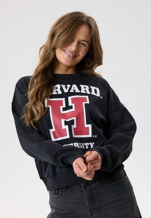 Jauna moteris su ilgais banguotais plaukais šypsosi, dėvi juodą Harvardo universiteto džemperį ir tamsias džinsines kelnes, stovėdama priešais paprastą šviesų foną.