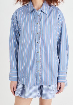 Person trägt ein blaues Hemd mit Knöpfen und vertikalen Streifen über einem blau-weiß karierten Rock oder Shorts.