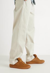 Pantaloni beige a gamba larga in cotone con dettagli sulle cuciture laterali, abbinati a sneaker in suede marrone con una suola in gomma spessa.