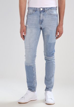 Mann trägt hellblaue Slim-Fit-Jeans mit Patch-Details und weiße Sneaker, steht vor einem schlichten hellen Hintergrund.