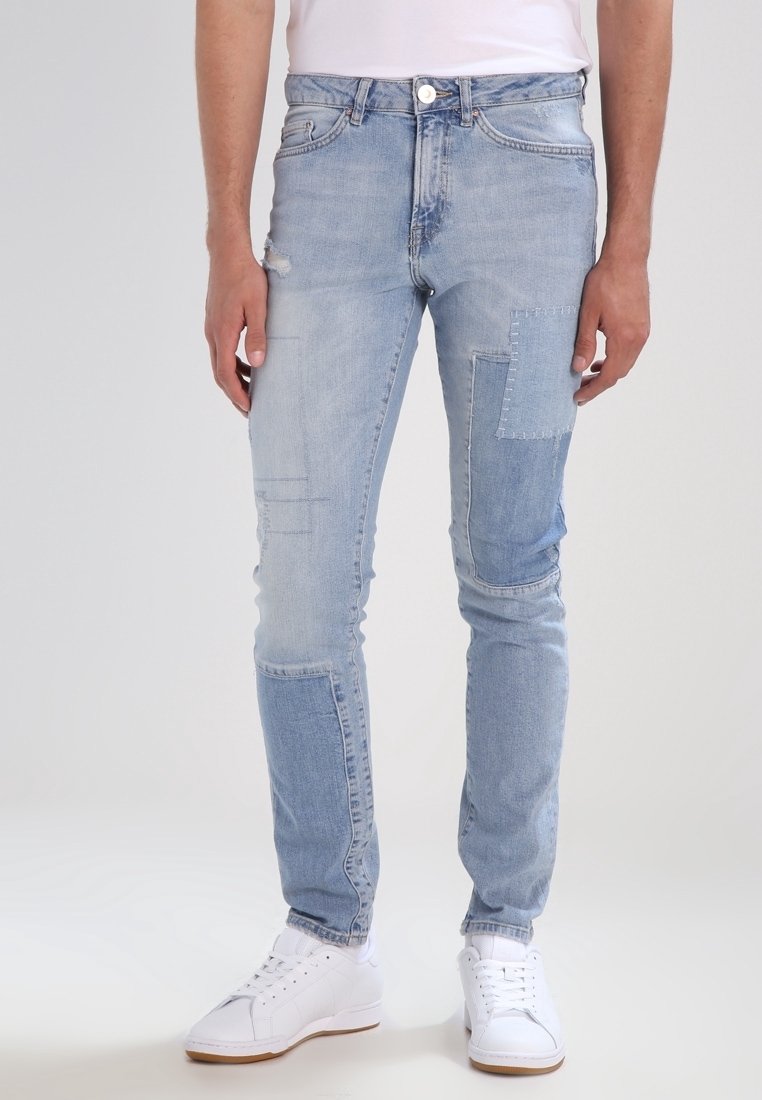 Homme portant un jean slim bleu clair avec des détails en patchs et des baskets blanches, debout devant un fond clair uni.