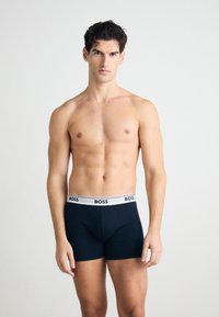BOSS temno modri bokseric z belim elastičnim pasom, na katerem je logotip blagovne znamke. Gladek material s prilegajočim oblikovanjem. Model stoji obrnjen naprej.