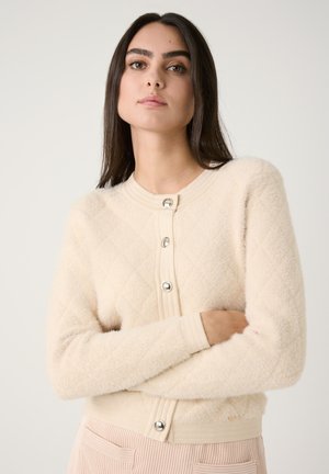 Gilet en tricot beige avec un motif en losanges, un col rond et des boutons en argent. Il a des poignets côtelés et une finition douce et texturée.