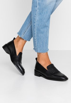Person, der hellblaue Jeans mit ausgefranstem Saum und schwarze Leder-Loafer mit geflochtenen Details auf weißem Boden trägt.