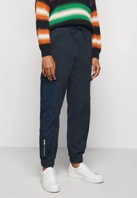 Marinblå cargobyxor med elastiska ärmslut, utrustade med en sidoficka med dragkedja och ett texturerat kontrastpanel i en ljusare blå färg på vänster ben.