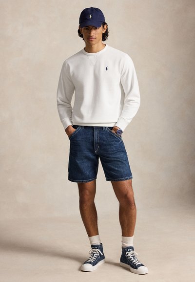 Homme portant une casquette bleu marine, un sweat-shirt blanc, un short en jean, des chaussettes blanches et des baskets montantes bleues, debout les mains dans les poches.