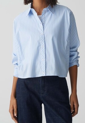 Light blue button-up shirt with white vertical stripes, cropped design, long sleeves rolled up, paired with dark denim pants.