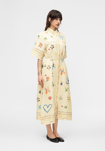 Μια γυναίκα στέκεται φορώντας ένα κρεμ midi πουκάμισο-φόρεμα με πολύχρωμα, χειροποίητα σχέδια λουλουδιών και καρδιών, με ζώνη δεμένη στη μέση και μαύρες μπαλαρίνες.