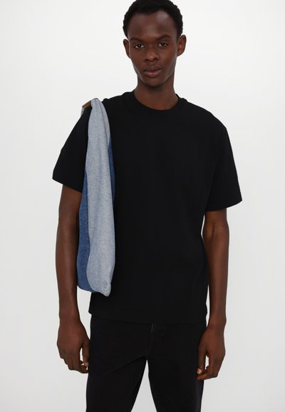 Tricou negru cu mânecă scurtă, purtat cu pantaloni închisi la culoare, având o geantă din denim aruncată pe un umăr, cu un design din denim deschis și închis.