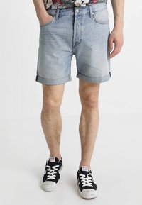 Shorts de mezclilla azul claro con dobladillos enrollados y cinco bolsillos, llevados con zapatillas de lona negras que tienen suelas de goma blanca y un logotipo.