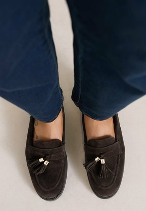 Paire de mocassins en daim marron foncé avec des glands portés avec un jean bleu sur un sol clair.