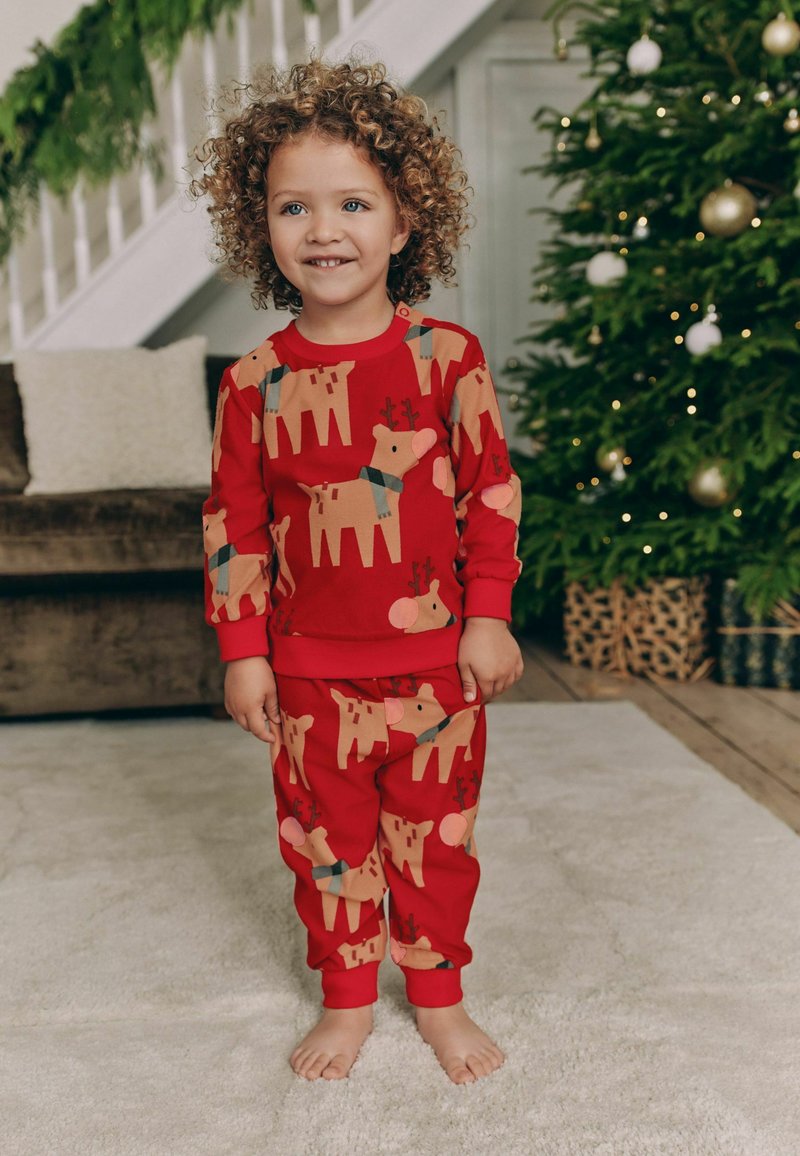 Punainen pyjamapaketti, jossa on leikkisiä porokuvioita, valmistettu pehmeästä kankaasta ja jossa on joustavat hihansuut. Taustalla näkyy joulukuusi.