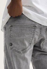 Graue Jeans aus Denim mit strukturierter Oberfläche, ausgestattet mit zwei Gesäßtaschen und einem schwarzen Etikett. Die Jeans werden mit einem weißen Hemd kombiniert.