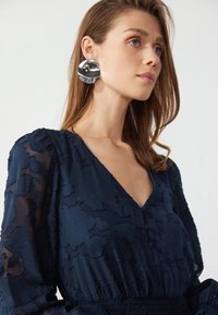 Robe marine avec broderies florales, manches longues transparentes, décolleté en V et taille smockée. Boucles d'oreilles circulaires en argent ajoutent une brillance contrastante.
