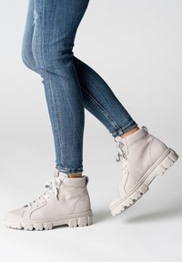 Baskets montantes en cuir gris clair avec lacets blancs, finition texturée et semelle épaisse. Portées avec un jean bleu ajusté.