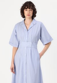 Abito camicia azzurro chiaro con strisce verticali bianche, maniche corte, vita aderente, tasche laterali e una silhouette rilassata.