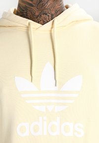 Žlutá mikina s kapucí vyrobená z bavlny, s bílým logem Adidas a třemi pruhy na přední straně, s tkaničkami a texturovaným povrchem.