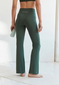Frau steht barfuß auf einer Yogamatte, trägt dunkelgrüne ausgestellte Leggings und Sport-BH, hält eine hellgrüne Wasserflasche in der linken Hand.