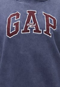 Μπλε φούτερ με μεγάλα κόκκινα γράμματα "GAP" περιγραμμισμένα με λευκό και τη λέξη "Original" σε μικρότερο λευκό γράψιμο μέσα στο γράμμα Α.