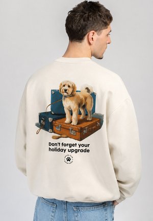 Mand iført cremefarvet sweatshirt med grafik af hund, der står på kufferter og tekst "Don't forget your holiday upgrade."