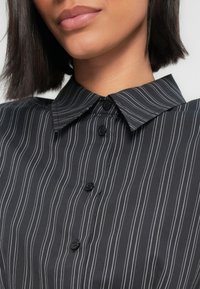 Κοντινό πλάνο του κάτω μέρους του προσώπου και του λαιμού που φοράει μαύρο πουκάμισο με λευκές κάθετες ρίγες και μαύρα κουμπιά, με μυτερό γιακά.