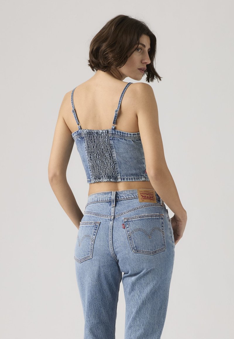 Donna che indossa un top corto in denim blu con spalline sottili e jeans in denim blu a vita alta, in piedi con la schiena rivolta verso lo spettatore e la testa leggermente girata a destra.