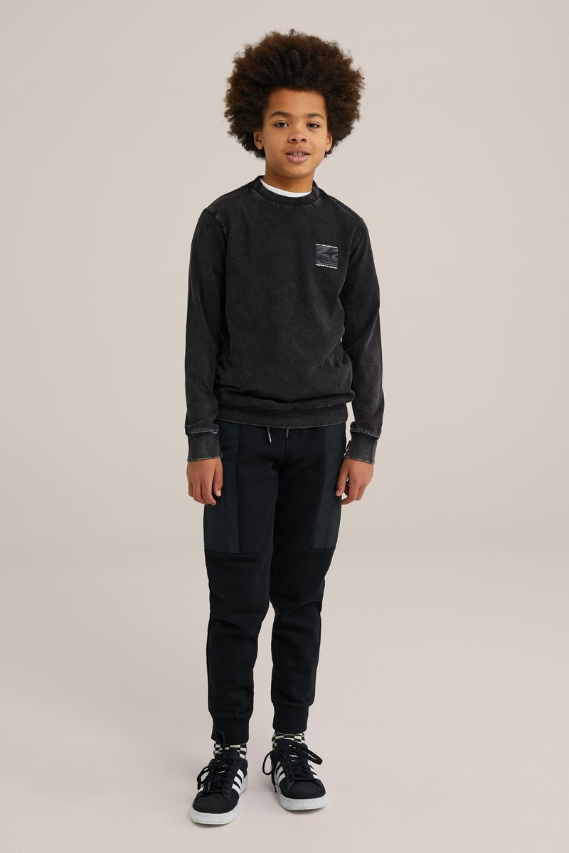 Schwarzes Langarm-Sweatshirt mit strukturiertem Taschen, kombiniert mit schwarzen Jogginghosen mit seitlichen Einsätzen. Schwarze Turnschuhe runden das Outfit ab.