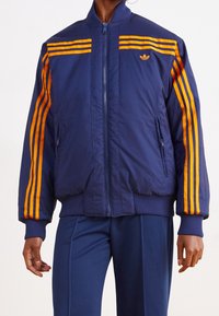 Blue bomber jacket with orange stripes, zippered front, ribbed cuffs and hem, featuring logo on chest and two side pockets.