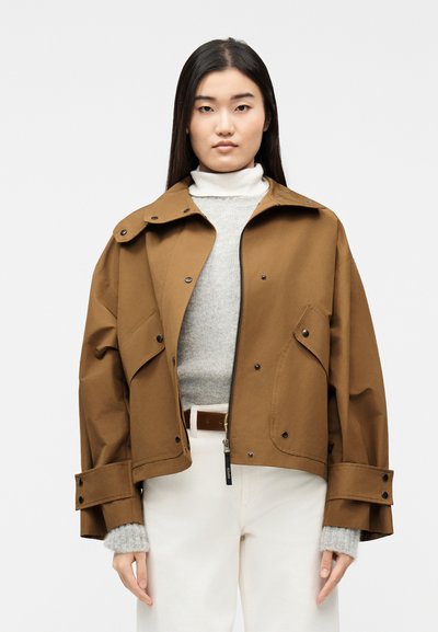Femme portant une veste marron surdimensionnée par-dessus un pull gris et un pantalon blanc, debout devant un fond blanc uni.