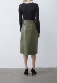 Maglietta nera a maniche lunghe abbinata a una gonna midi in pelle verde oliva e tacchi a spillo neri, che mostrano una silhouette elegante e aderente.